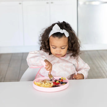 A toddler enjoys scrambled eggs and berries on a pink silicone grip plate by Bumkins, sitting in a white kitchen.