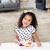 A young girl smiles, wearing a heart-patterned shirt, holding a fork over her Bumkins Silicone Grip Plate in pink.