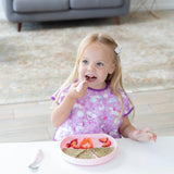 A young girl enjoys sliced fruits and toast on a Bumkins Silicone Grip Plate in pink, wearing a purple bib with cute designs.