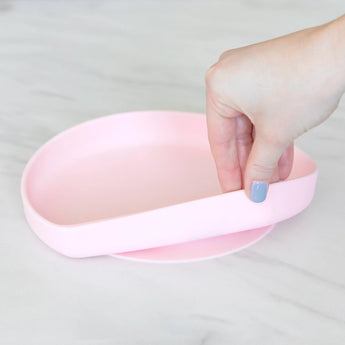 A hand lifts a Bumkins Pink Silicone Grip Plate, divided with sections, on a gray marble surface. Food-safe silicone used.