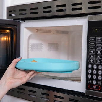 A hand places the Bumkins Silicone Grip Tray: Blue into the microwave, revealing the control panel and interior.