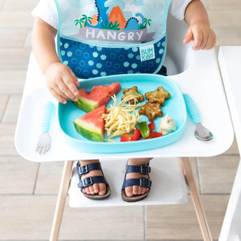 A toddler enjoys a meal from a Bumkins Silicone Grip Tray: Blue, featuring watermelon, noodles, and star-shaped treats.