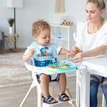 A toddler in a high chair enjoys noodles from a Bumkins Silicone Grip Tray: Blue, wearing a bib, as a woman nearby holds a fork of food.