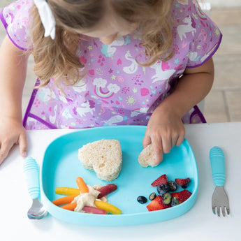 A child in a unicorn dress enjoys heart-shaped sandwiches with fruits and veggies on a Bumkins Silicone Grip Tray: Blue.