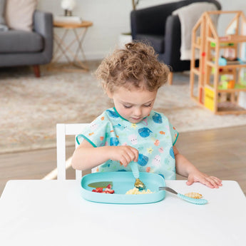 A toddler with curly hair eats from a blue Bumkins Silicone Grip Tray at a white table, toys and a sofa in the background.