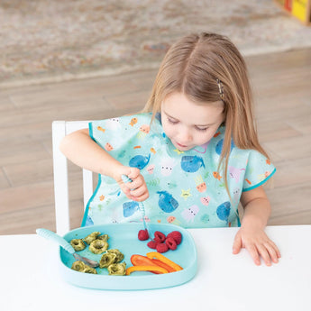 A child in a colorful bib enjoys raspberries, pasta, and peppers with a fork on a stylish Bumkins Silicone Grip Tray: Blue.