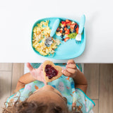 Child eating toast with jam on a Blue Silicone Grip Tray by Bumkins, with a plate of scrambled eggs and fruit at the table.