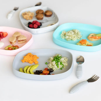 Four gray Silicone Grip Trays by Bumkins, arranged with various foods and utensils on a white surface.