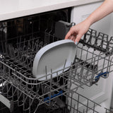 Placing the Bumkins Silicone Grip Tray, gray with strong suction base, into an open dishwasher against a white countertop.