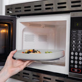 A hand places a white plate of food into a microwave on Bumkins Silicone Grip Tray: Gray, crafted from food-safe silicone.