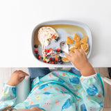 Child enjoys pancakes with fruit and yogurt on a Bumkins Silicone Grip Tray: Gray, wearing sea creature bib. Top view.