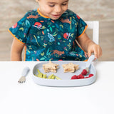 A child in a colorful bib sits at a Bumkins Silicone Grip Tray: Gray with star sandwiches, green snacks, raspberries, using cutlery.