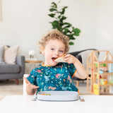 A curly-haired child enjoys a sandwich on a Bumkins Silicone Grip Tray, wearing a colorful bib. Nearby, a plant and wooden toy house add charm.