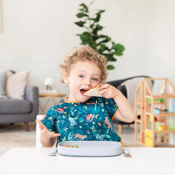 A curly-haired child enjoys a sandwich on a Bumkins Silicone Grip Tray, wearing a colorful bib. Nearby, a plant and wooden toy house add charm.