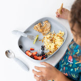 Child in colorful outfit enjoys meal with Bumkins Silicone Grip Tray (Gray), featuring eggs, fruit, and oat heart pancakes in silicone dishes.