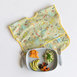 Baby bib with playful graphics, quinoa with peas, avocado slices, and mashed sweet potatoes on a Bumkins Silicone Grip Tray: Gray.