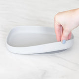 Hand holds Bumkins Silicone Grip Tray: Gray with sturdy suction base angled on white surface.