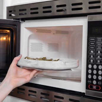 A person uses the Bumkins Silicone Grip Tray: Marble, made from food-safe silicone, to steadily place a plate of food into a microwave.