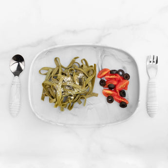 A Silicone Grip Tray: Marble by Bumkins holds spinach pasta with parmesan, cherry tomatoes, and olives, alongside a spoon and fork.