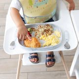 A toddler in a high chair enjoys breakfast using a Bumkins Happy Camper bib and Silicone Grip Tray: Marble for scrambled eggs and hash browns.