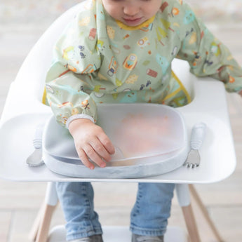 Toddler in a high chair with Bumkins Grip Dish, using colorful bib and utensils, reaching for food beside a Grip Tray.