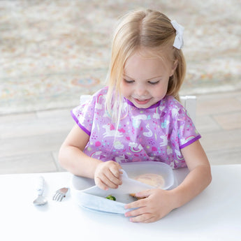 A young blonde child in a purple unicorn bib sits at a white table, eating from a Bumkins Silicone Grip Tray in Marble. The strong suction base keeps it steady as she holds a spoon, while utensils are set beside her.