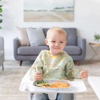 A toddler in a high chair enjoys food on a Bumkins Silicone Grip Tray: Marble. A gray couch and painting add charm to the cozy scene.