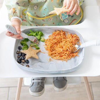 A child enjoys pasta, broccoli, and blueberries on Bumkins Marble Silicone Grip Tray with a secure suction base.