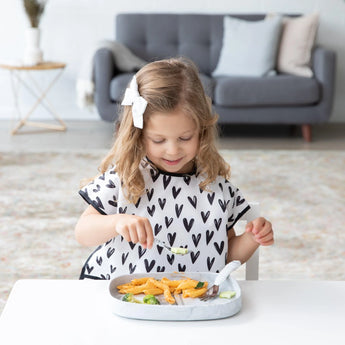 A girl in a heart-patterned shirt enjoys her meal on a Bumkins Silicone Grip Tray: Marble at a small table with a gray sofa behind her.