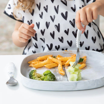 Child enjoying pasta, broccoli, and cucumber on a Bumkins Silicone Grip Tray: Marble. Wearing a white shirt with black hearts.