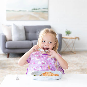 A little blonde girl happily eats with her purple bib and Bumkins Silicone Grip Tray in Marble, keeping mealtime mess-free.