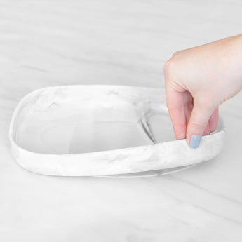 A hand grips the edge of a Bumkins Silicone Grip Tray: Marble on a marble surface.