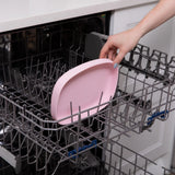 A person places a Bumkins Silicone Grip Tray: Pink into the open dishwasher rack.