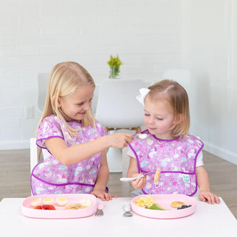 Two blonde kids enjoy meals with Bumkins Silicone Grip Tray: Pink; one uses a spoon to feed the other at the table.