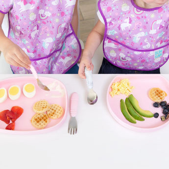 Two kids in unicorn bibs enjoy breakfast on a Bumkins Silicone Grip Tray: Pink with waffles, eggs, and fresh fruit.