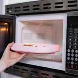 A hand places food on a Bumkins Silicone Grip Tray: Pink with a suction base inside the microwave, revealing the control panel on the right.