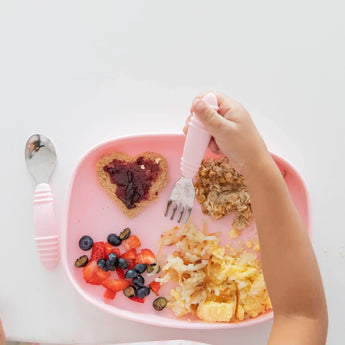 Childs hand holds fork over Bumkins Silicone Grip Tray: Pink, with heart-shaped toast, fruit, hash browns, and cereal; spoon nearby.