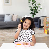 A curly-haired child with a star item sits with snacks on a Bumkins Silicone Grip Tray in pink, held securely by its suction base.