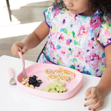 A child with a colorful bib sits at a table with a Bumkins Silicone Grip Tray: Pink, holding rolled sandwiches, cucumbers, and blueberries.