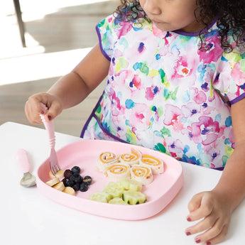 A child with a colorful bib sits at a table with a Bumkins Silicone Grip Tray: Pink, holding rolled sandwiches, cucumbers, and blueberries.