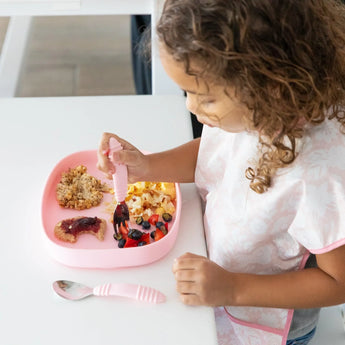 A child in a bib uses a fork to eat berries and oatmeal from the Bumkins Silicone Grip Tray: Pink at the white table.