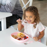 A girl with curly hair enjoys breakfast on a Bumkins Pink Silicone Grip Tray at a white table, filled with eggs, berries, and toast.