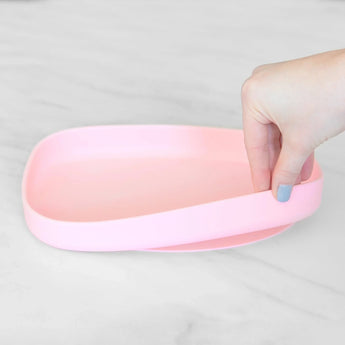 A hand with painted nails holds the corner of a pink Bumkins Silicone Grip Tray on a marble surface.