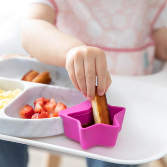 A child dips food into a star-shaped pink Bumkins Silicone Little Dipper on a tray with strawberries and more.