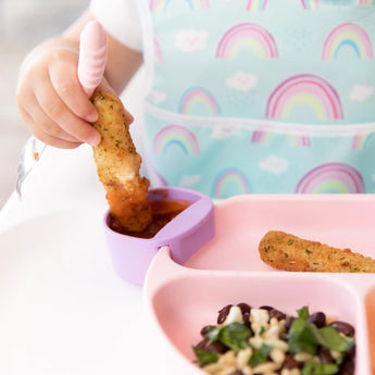 Child dips mozzarella stick into sauce on a pink tray with beans & rice, wearing a rainbow bib, using Bumkins Silicone Little Dippers.