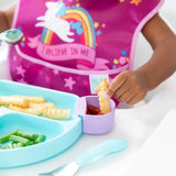 A child enjoys crinkle fries with ketchup in Bumkins Silicone Little Dipper, wearing a bib featuring a unicorn and rainbow design.