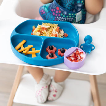 Child in a high chair with pasta, cheese sticks, and fruit on a blue plate. Yogurt in purple cup with Bumkins Silicone Little Dipper.