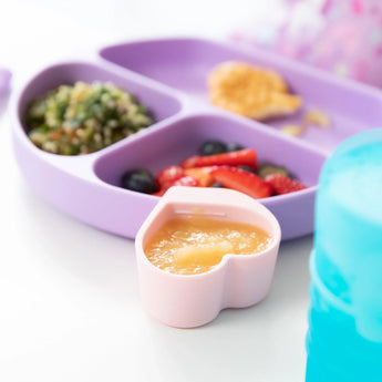 Purple divided plate with salad, heart-shaped cup with applesauce, and Bumkins Silicone Little Dipper in Lollipop on a white surface.