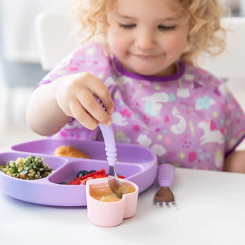 A curly-haired toddler in a purple bib eats with a Bumkins Silicone Little Dipper from a sectioned purple plate.