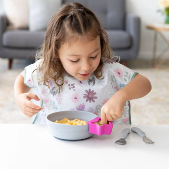 A child eats cereal with a pink Silicone Little Dipper by Bumkins at the table; two spoons are nearby.